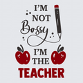 A graphic design with the text 'I'M NOT Bossy I'M THE TEACHER' in black and red lettering, accompanied by two red apples and a black-and-red pencil on a light gray background.